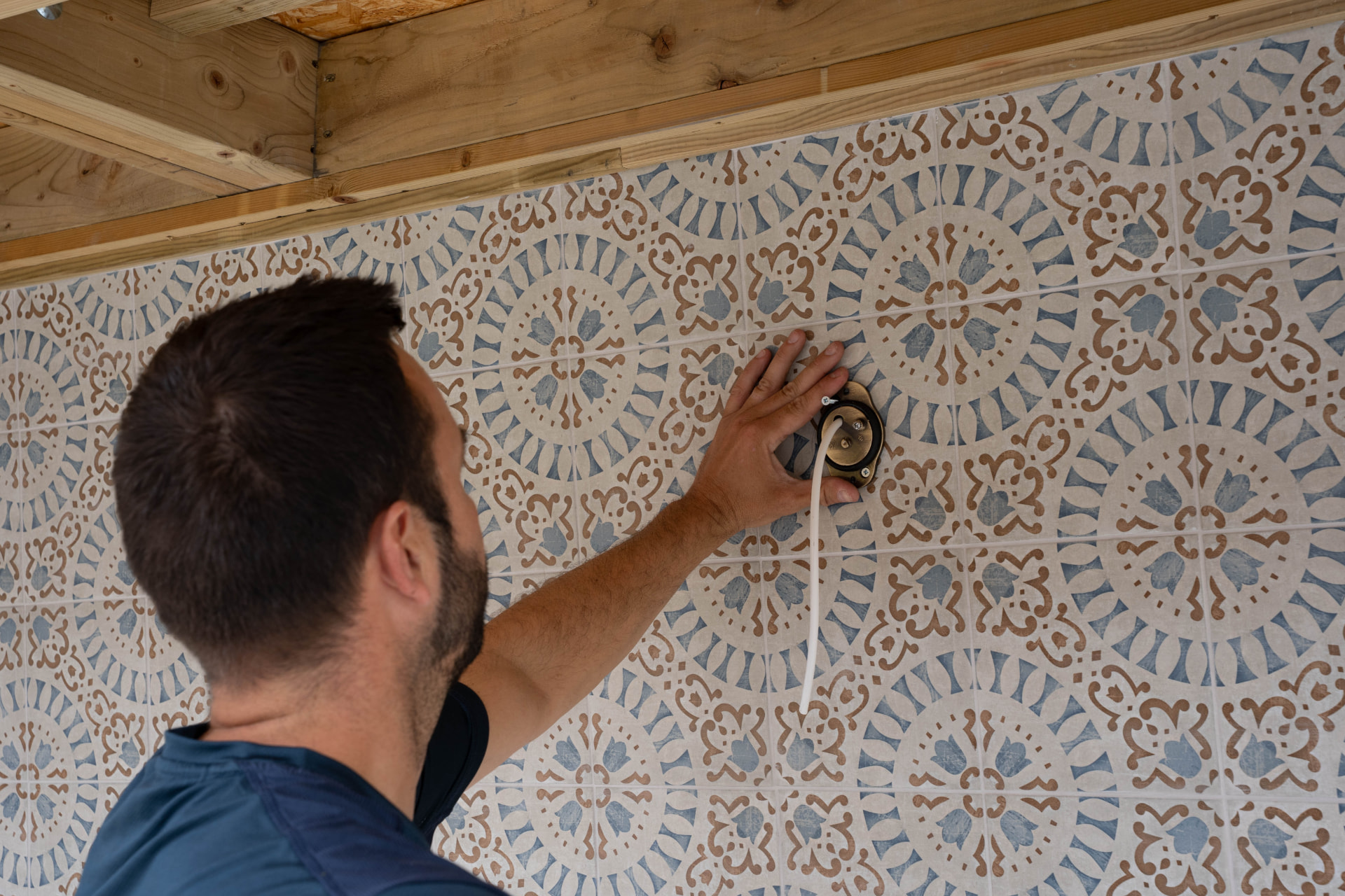 Electrician fitting an exterior wall light point onto a patterned tiled wall under a timber canopy